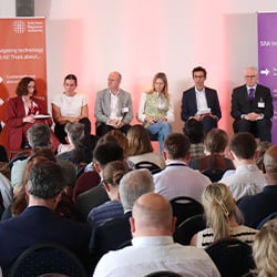 A panel of six people is seated on a stage at a conference. They are facing an audience. The backdrop features text related to navigating regulatory challenges and technology. Audience members are visible in the foreground.